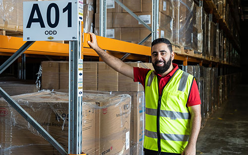 A staff member wearing a high‑visibility vest stands beside large boxed inventory on warehouse shelving.