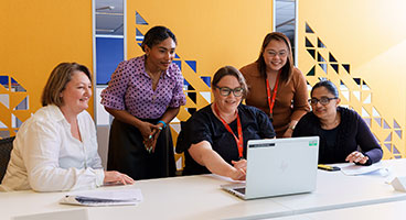 Five people gather around a laptop at a meeting table, collaborating in a modern office with patterned yellow wall panels.
