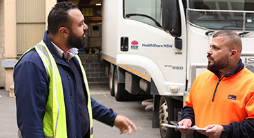 A man standing with a clipboard at a hospital dock, chatting to a colleague with the HealthShare NSW truck in the background