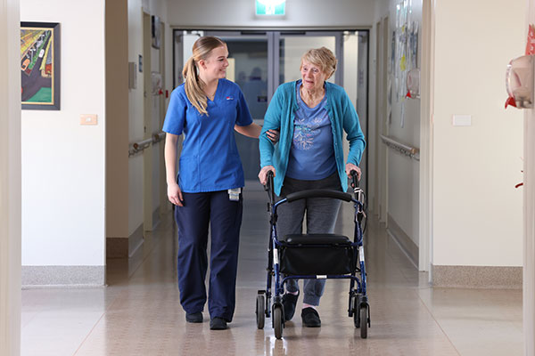 A staff member walks beside a person using a walker in a hallway of a care facility.