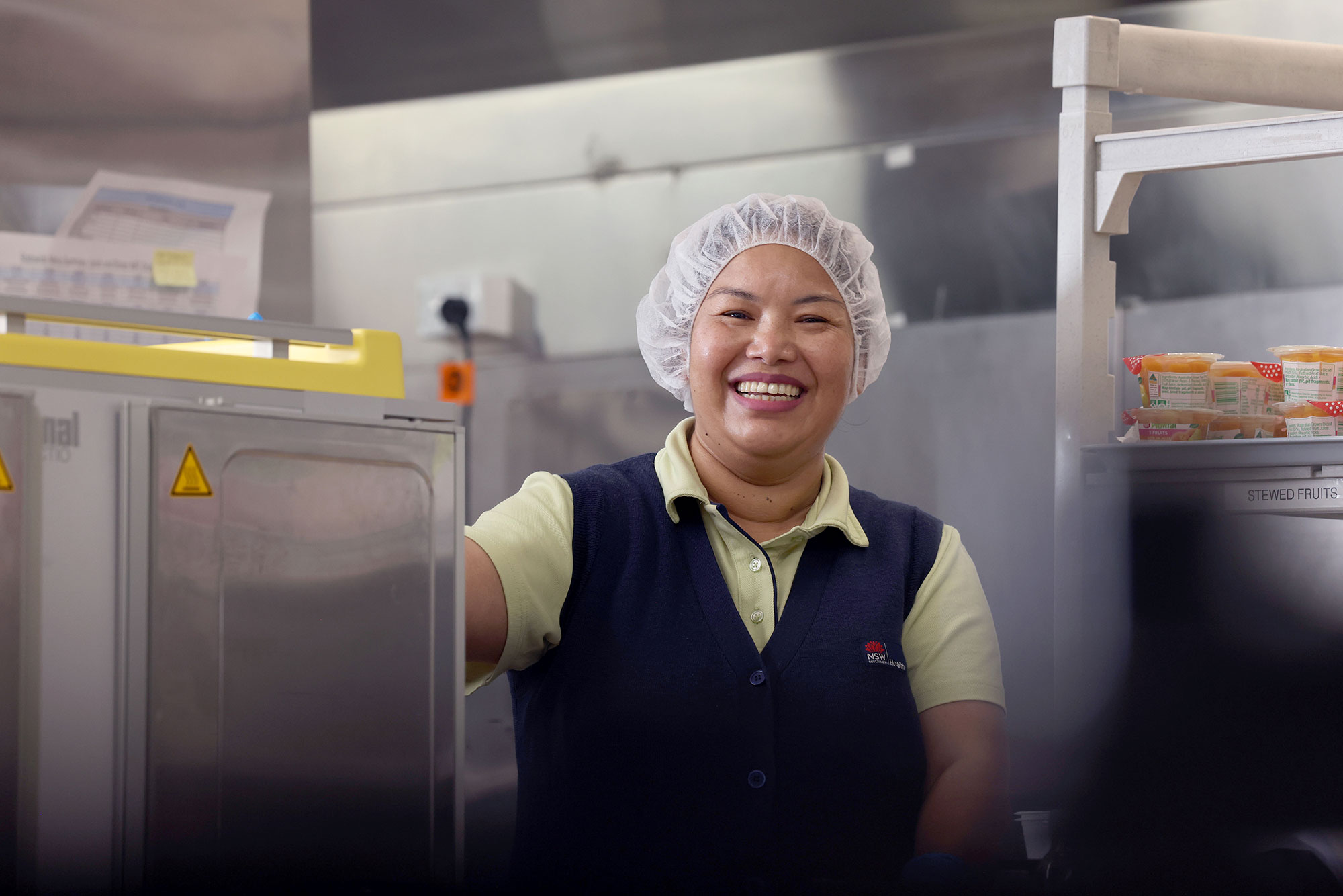 A staff member wearing a hairnet and uniform stands in a food‑service area next to heating equipment and shelves of packaged items.